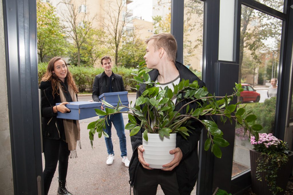 Kaksi miestä ja nainen kantavat tavaroita kerrostalon alaovesta sisään.