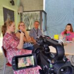 Etualalalla videokamera. Taustalla pöydän ääressä aikuisia haastateltavia ja nuori juontaja.